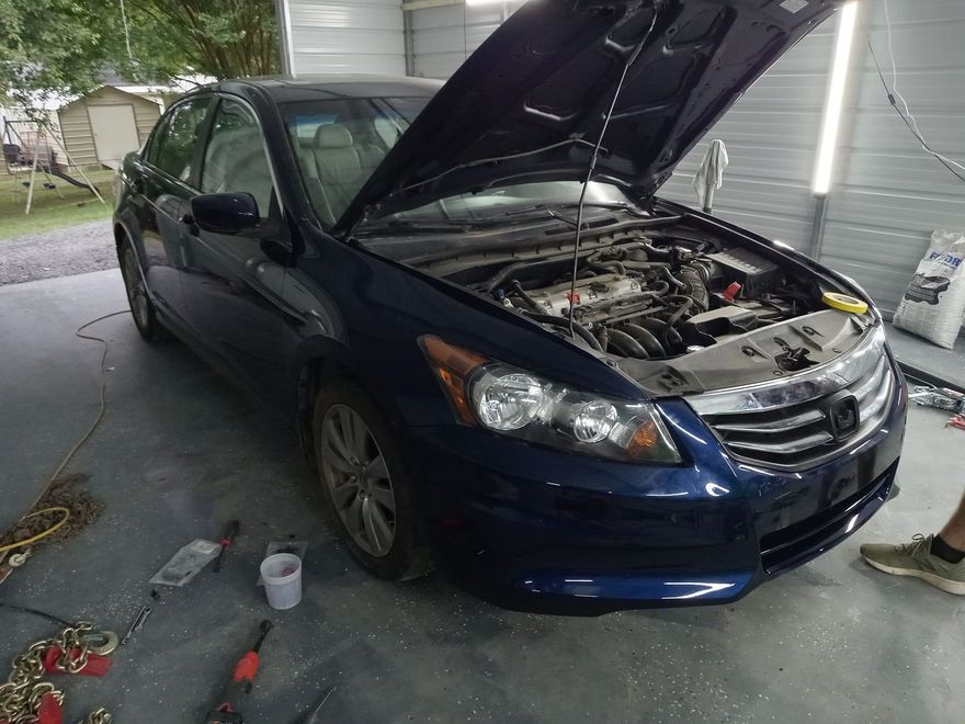 A dark blue sedan with its hood open, parked inside a garage workshop with a person's feet visible on the right.