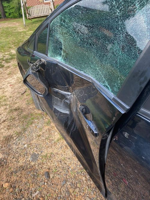 A black car with a severely dented side door and shattered window parked on a gravel surface.