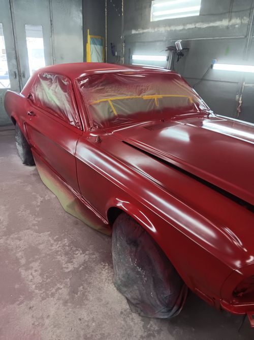 A classic red Ford Mustang being painted in a professional spray booth with windows masked off.