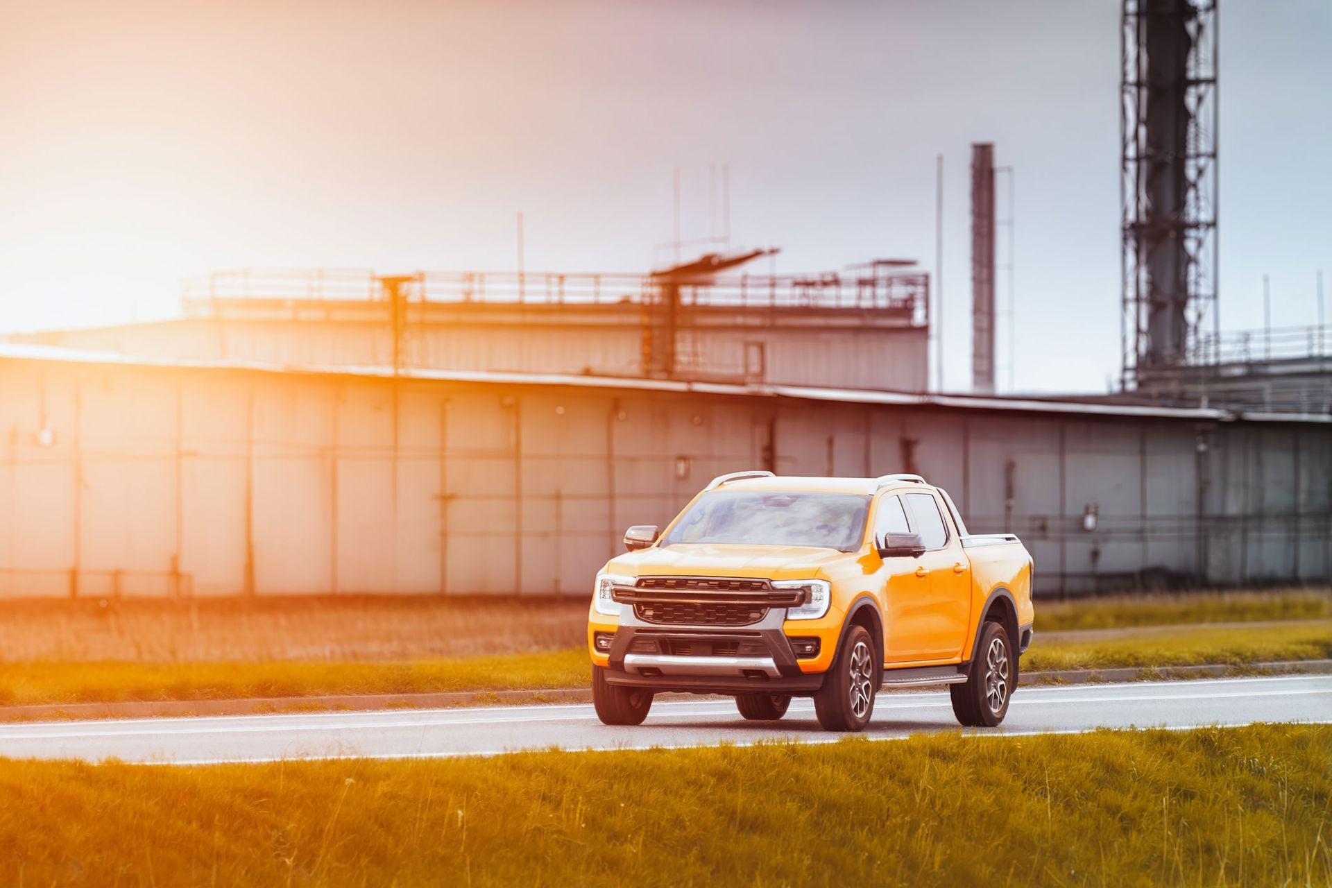 Yellow pickup truck driving on a road near industrial buildings in sunlight.