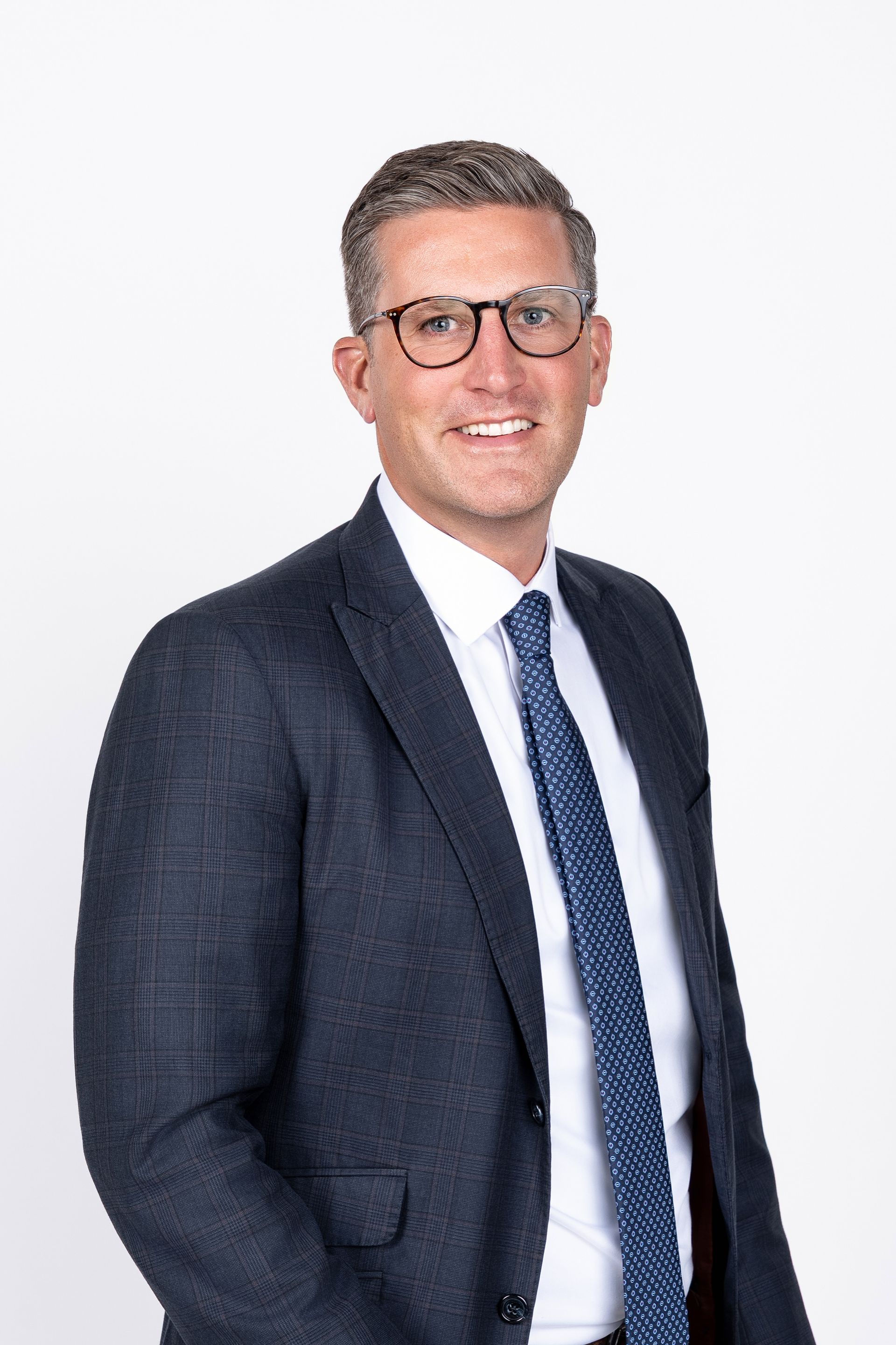 A man in a suit and white shirt is smiling in front of a brick wall.