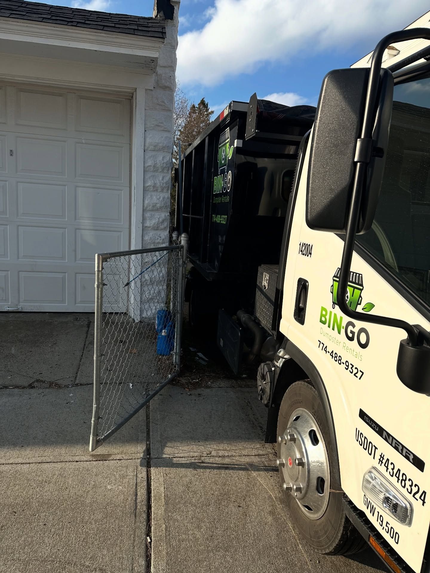 A beige Bingo waste removal truck parked in a residential driveway, partially obscuring an open chain-link fence gate.