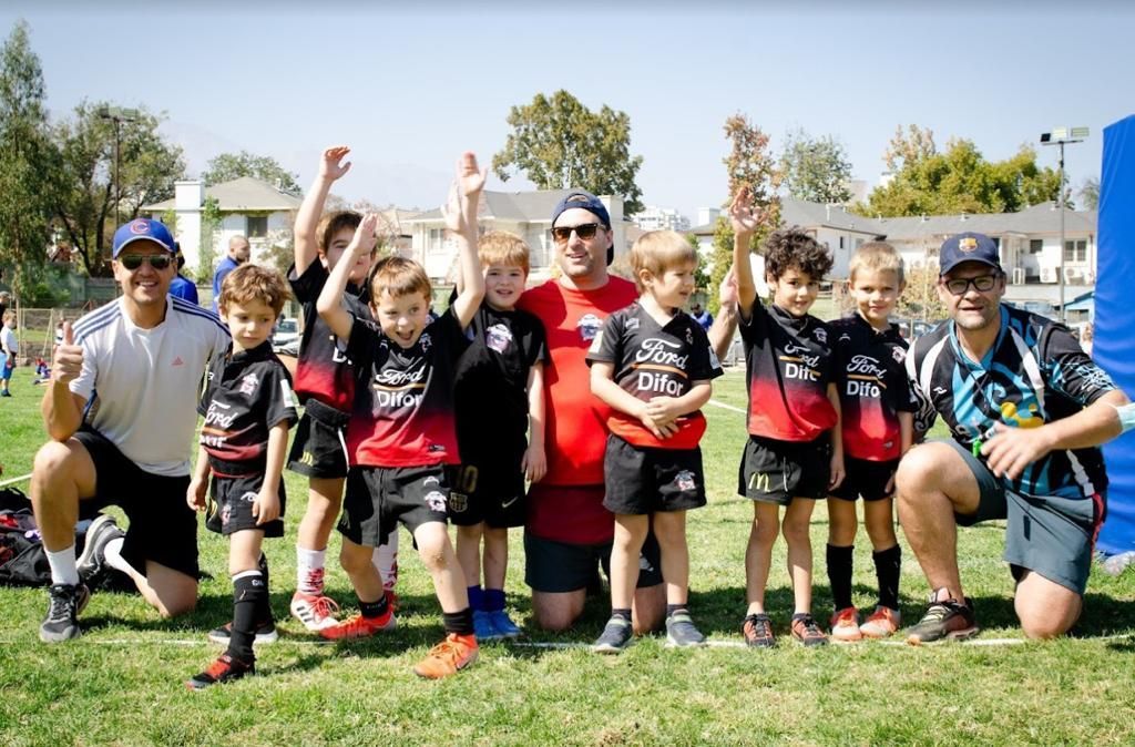 El equipo de rugby juvenil posa en un campo de césped, levantando las manos, con los entrenadores en un clima soleado.