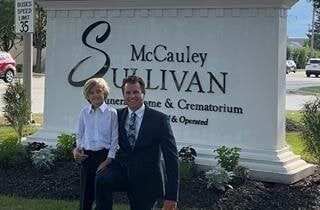 Two people pose in front of a funeral home sign. A man kneels next to a person in a white shirt. The sign is white with black text.
