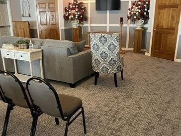 Interior of a waiting room with floral arrangements, a sofa, chairs, and a small table.