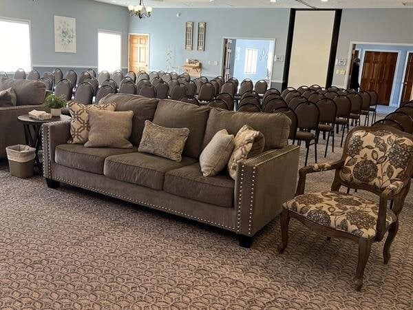A gray sofa and patterned chair in a large room set up for an event, rows of chairs in the background.