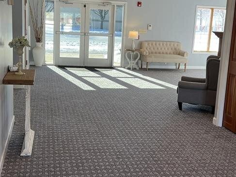 Bright lobby with a neutral carpet, glass doors, a couch, armchair, and a side table. Sunlight streams in.