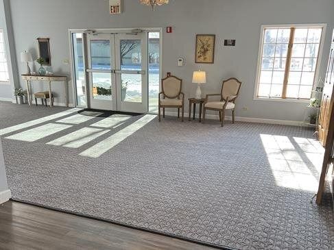 A bright room with carpet, two chairs, and an entrance with glass doors; sunlight casts shadows.