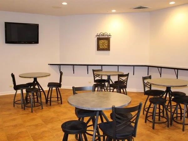 Interior with round tables, black stools, a mounted TV, and a wall shelf. Beige tile floor.