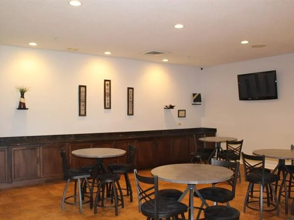 Empty lounge area with round tables, black stools, a bar, wall art, and a mounted TV.