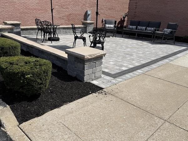 Outdoor patio with seating, brick retaining wall, and shrubs. Concrete sidewalk in foreground.