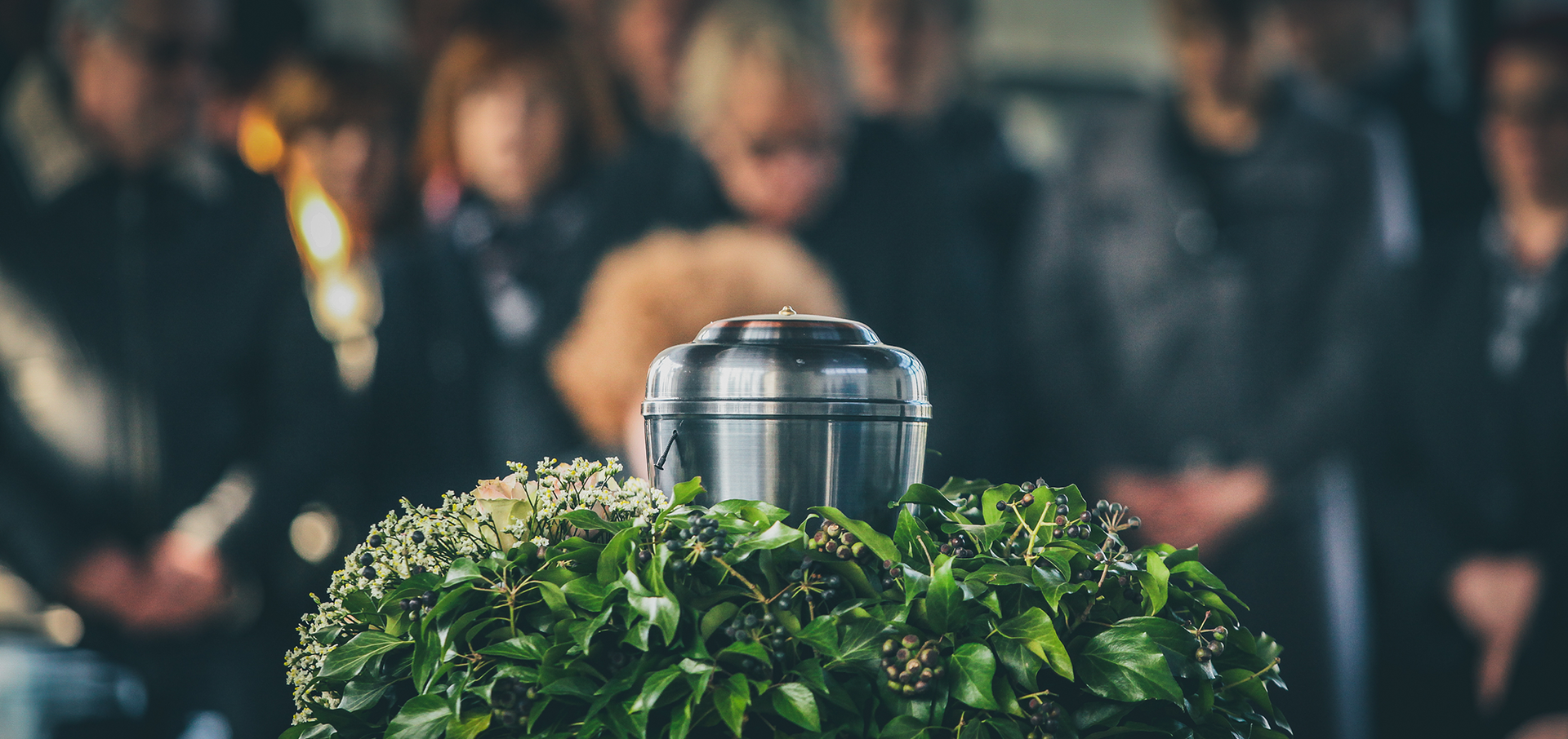Silver urn on a bed of green leaves, surrounded by people in dark clothing, at a somber gathering.
