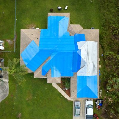 Overhead view of house with blue and white tarps covering damaged roof. Cars parked in driveway.