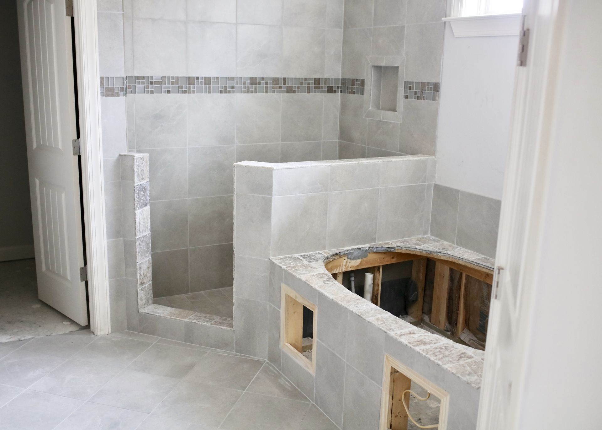 Bathroom with gray tiled shower stall, partially constructed tub area, and open doorway.