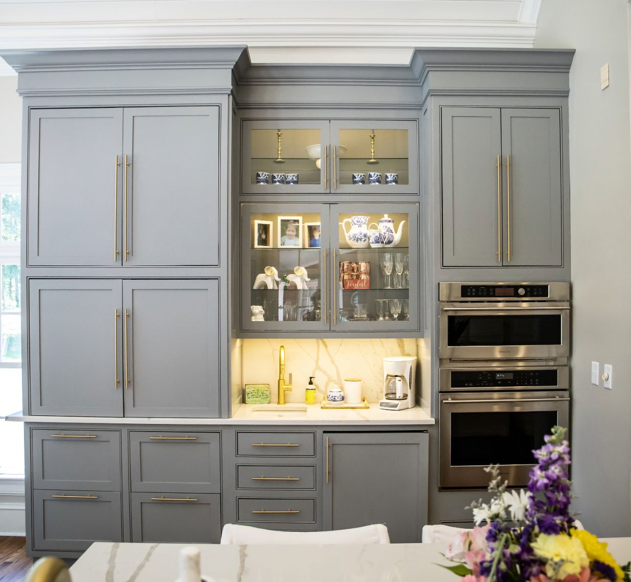 Gray kitchen cabinetry with built-in refrigerator, ovens, glass-front display, and counter with coffee maker.