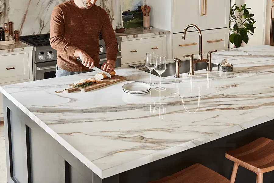 Man cutting food on a marble countertop in a modern kitchen, with black island and wooden stools.