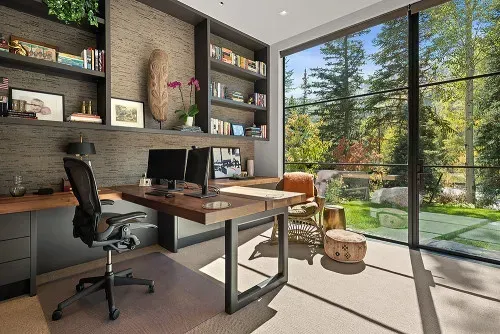Home office with desk, built-in shelves, large windows, and views of lush greenery.