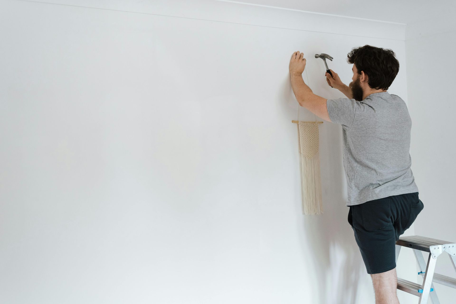 Man on a ladder hammering a nail into a white wall; beige paper strip hangs below.