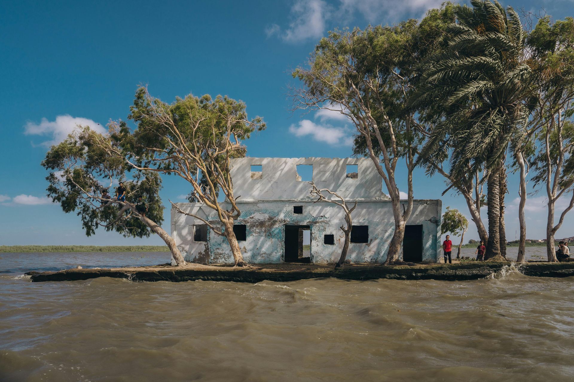 Dilapidated white building partially submerged in water; trees surround it. Blue sky, rough water.