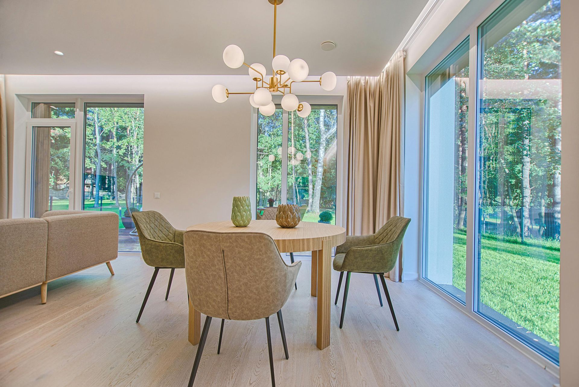 Dining room with round table, green chairs, gold chandelier, and large windows overlooking trees.