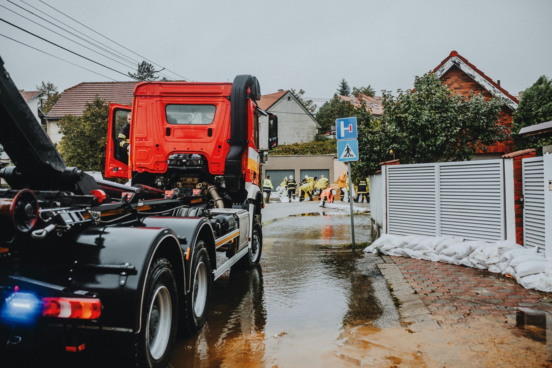 Red fire truck in a flooded street. Emergency responders in yellow vests work near sandbags protecting a house.