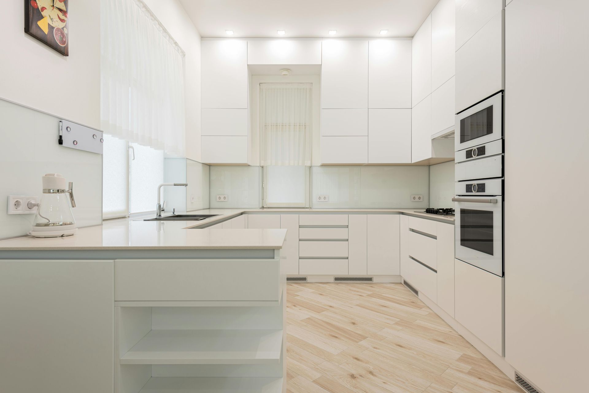 Modern, all-white kitchen with minimalist cabinets, built-in appliances, and light wood flooring.