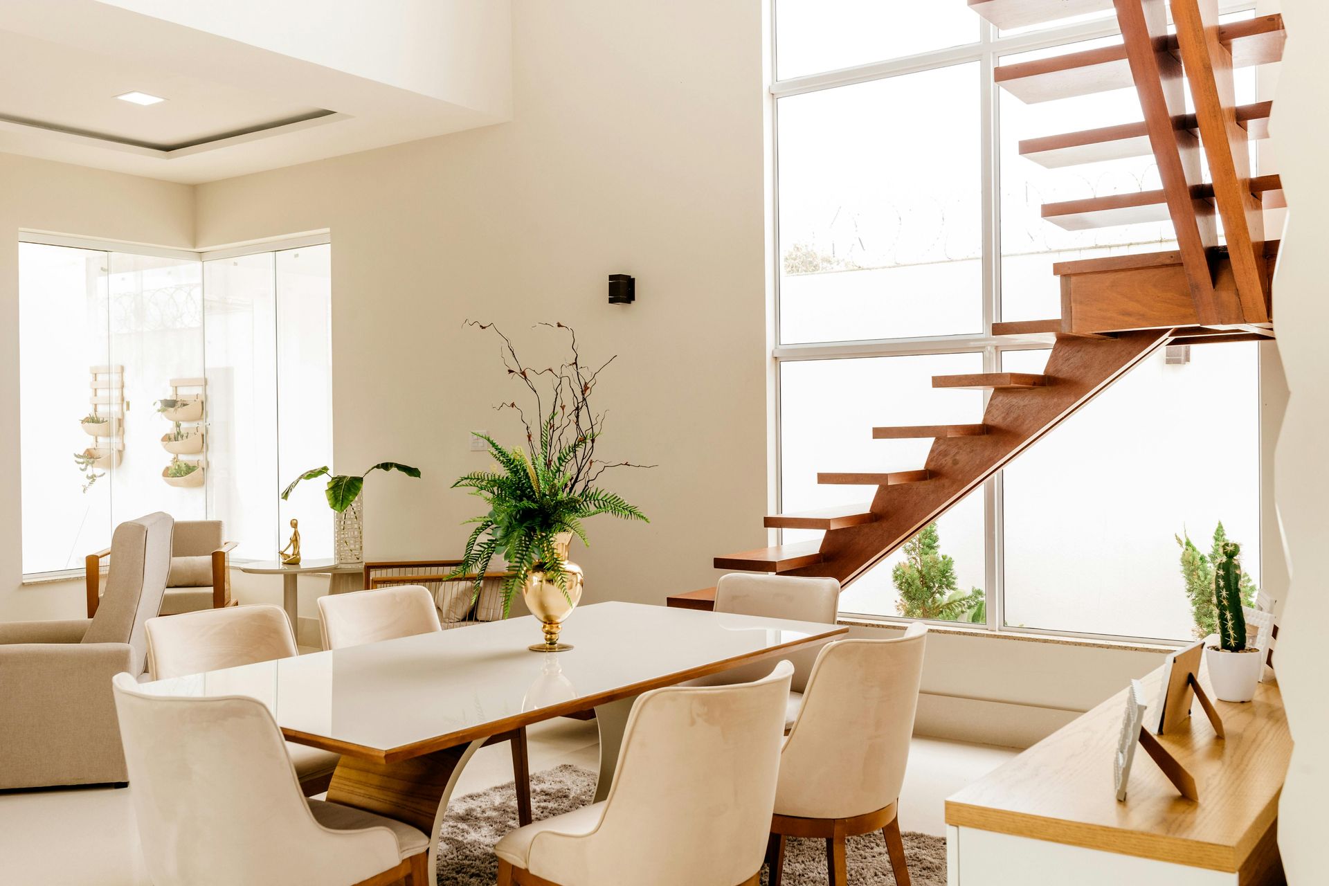 Bright dining area with white table and chairs, wooden staircase, large windows, and tall floral arrangement.