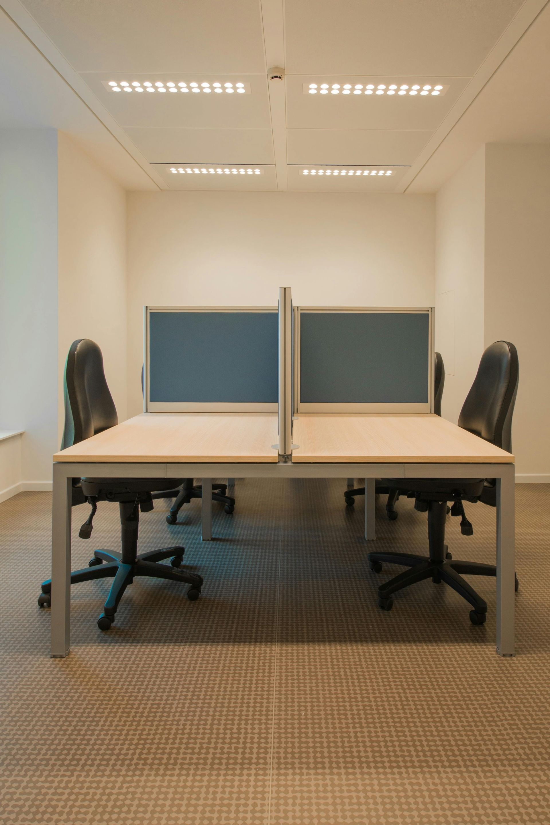 Office cubicles with desks, chairs, and blue partitions, in a brightly lit room.