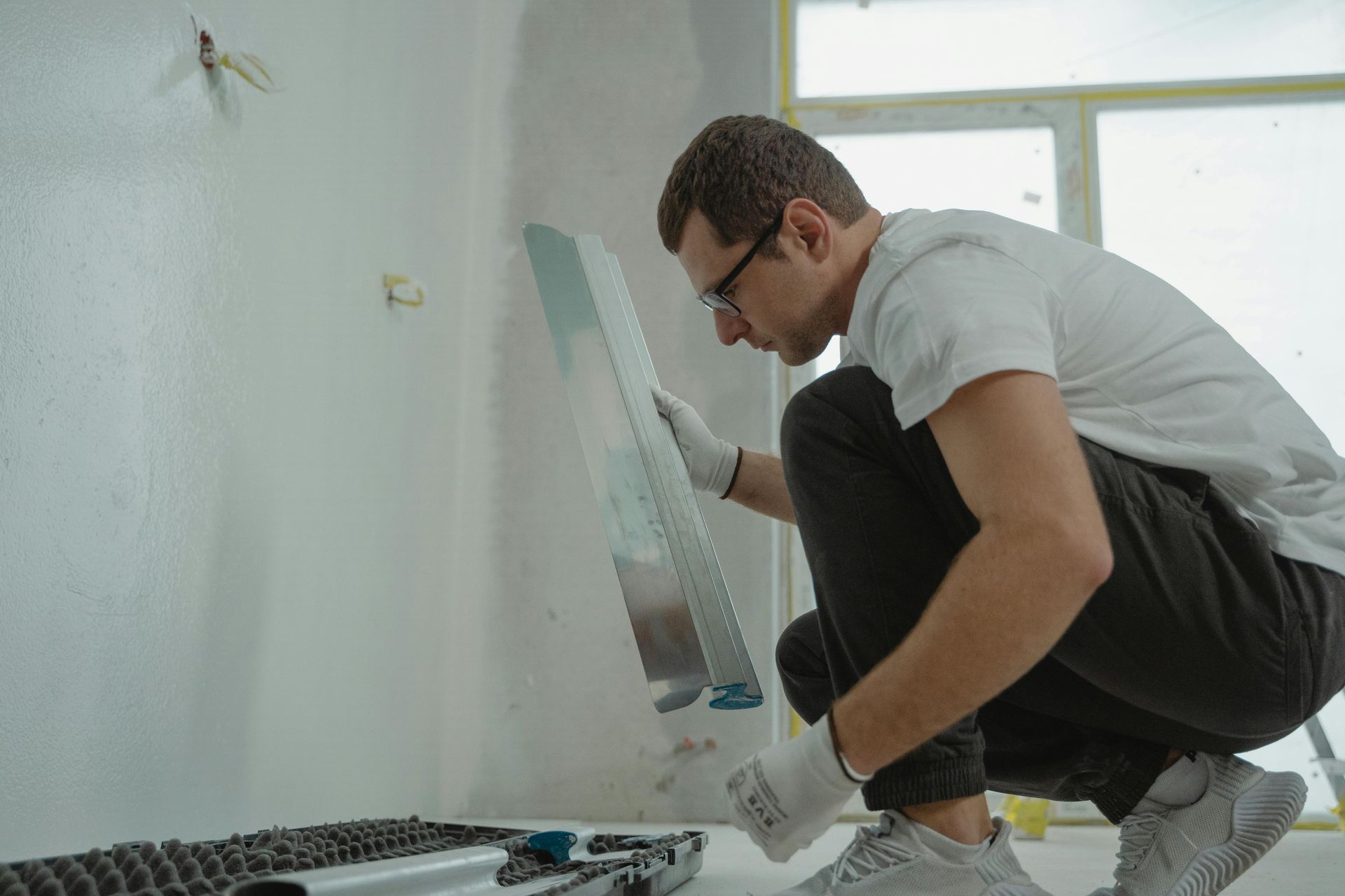 Man in glasses smoothing plaster on wall with trowel. Indoors, white wall and window.