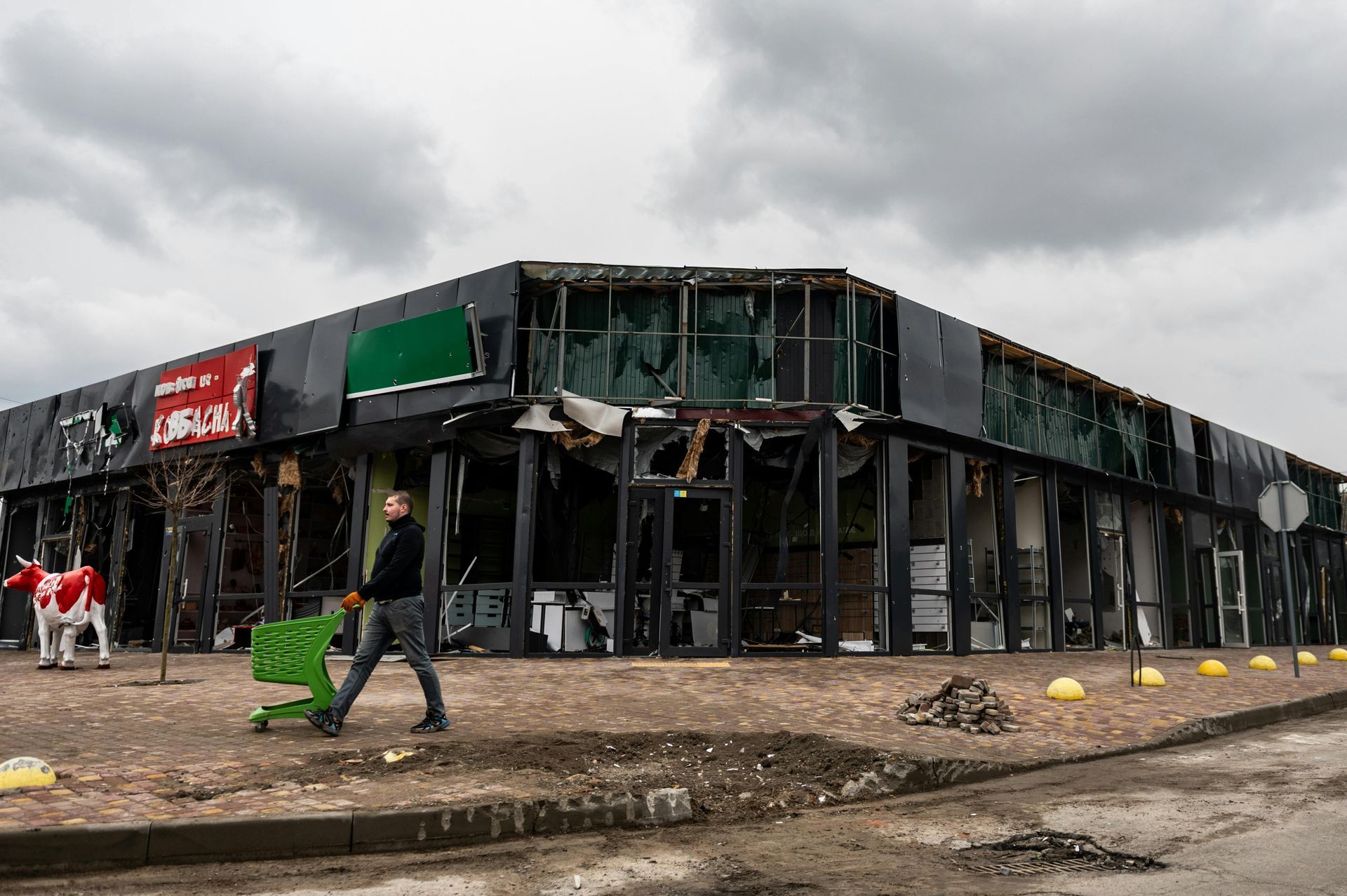 A person walks past a destroyed commercial building with a cow statue, under a cloudy sky.
