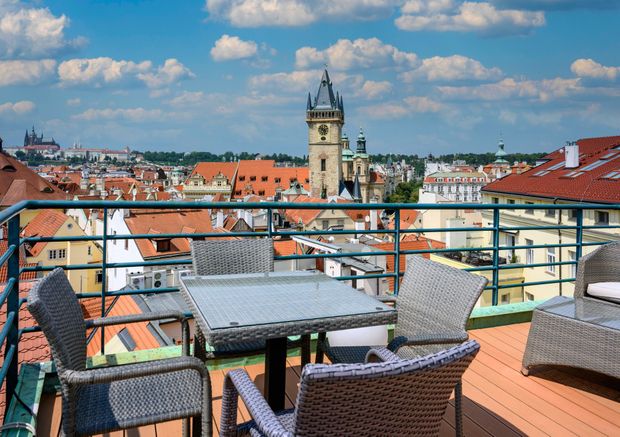 Rooftop terrace overlooking Prague's Old Town Square with seating for a meal or drinks. Blue sky with clouds.