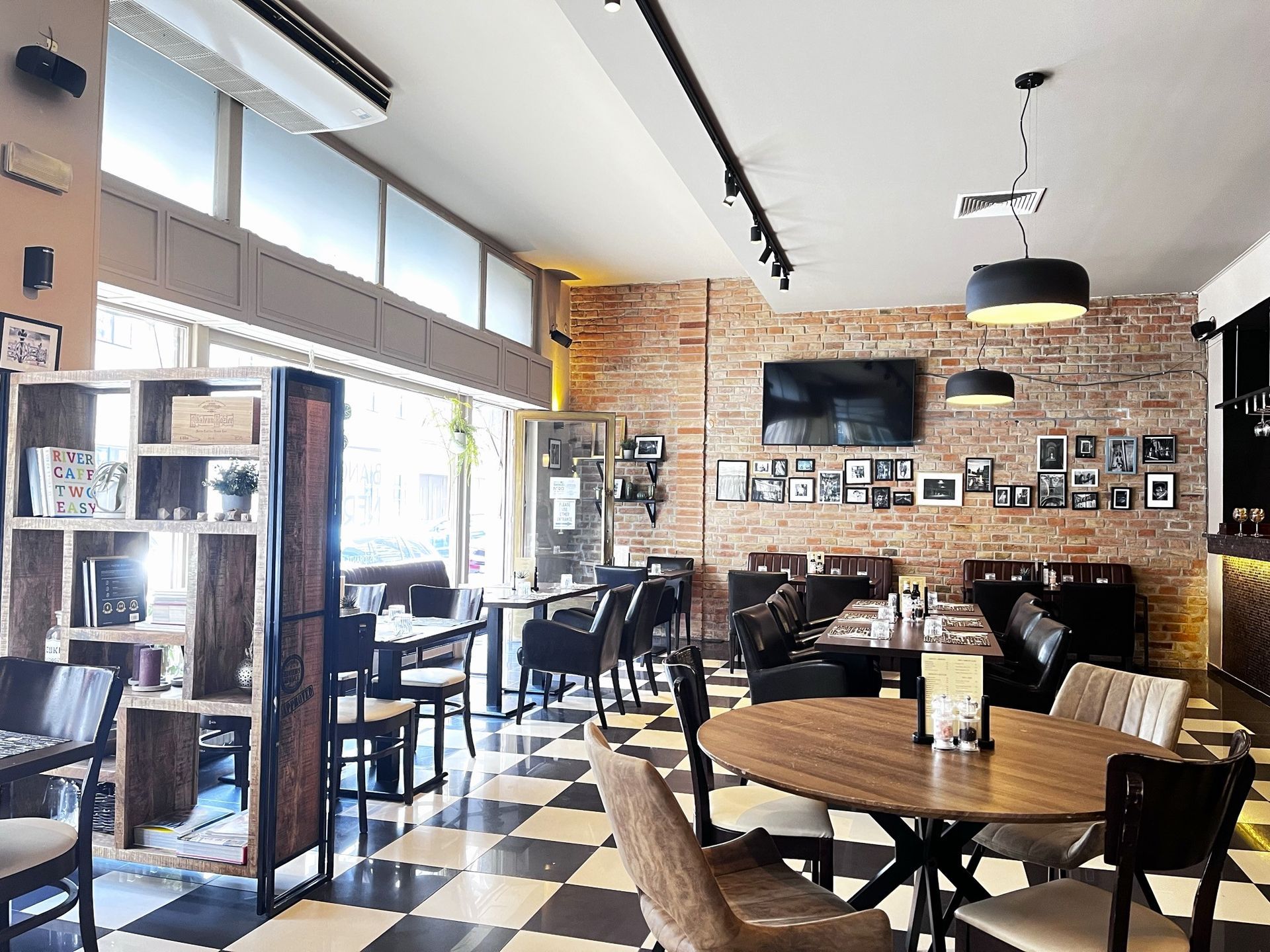 Restaurant interior with checkered floor, brick wall, tables, chairs, and decorative shelving.