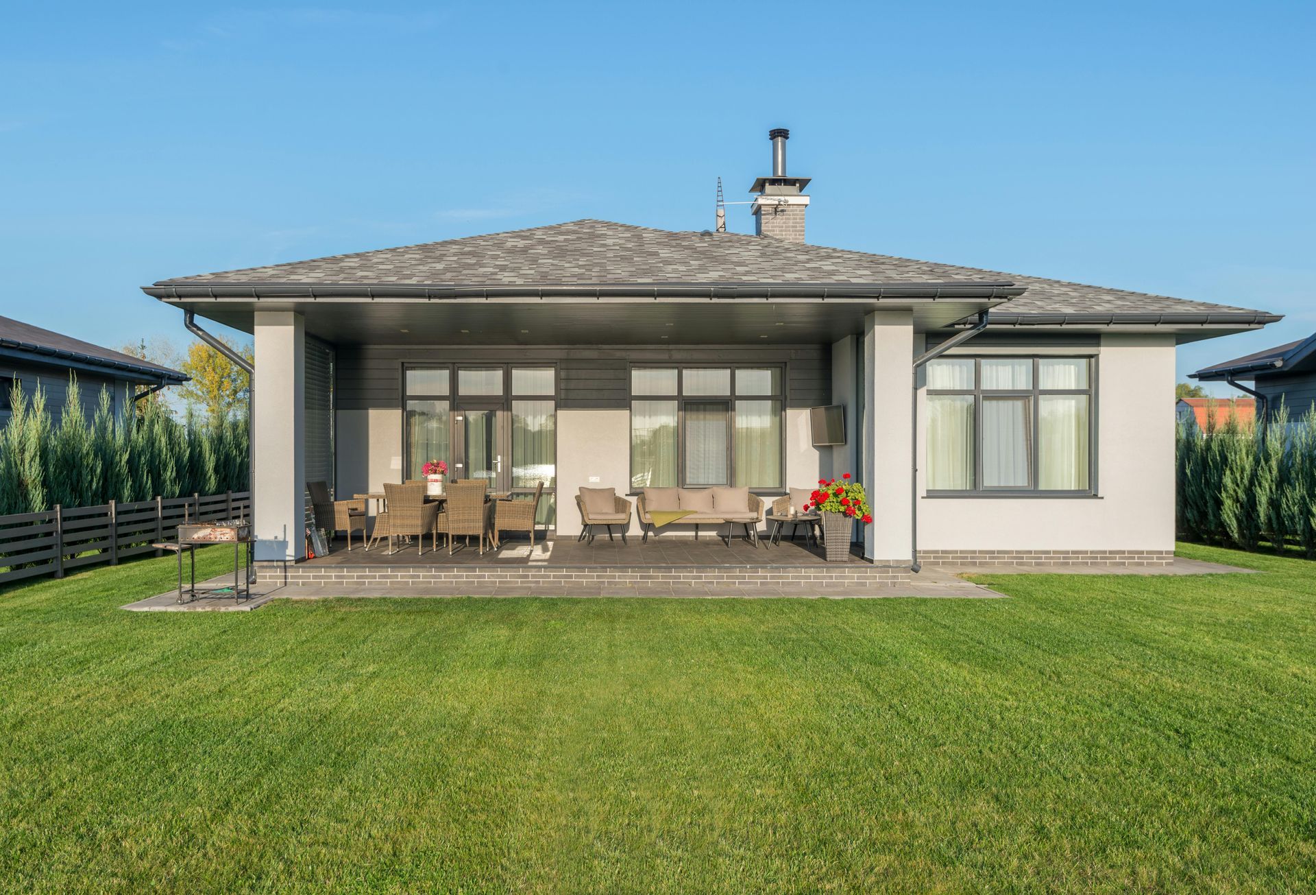 A modern, light-colored single-story house with a covered patio, outdoor seating, and a large green lawn under a blue sky.
