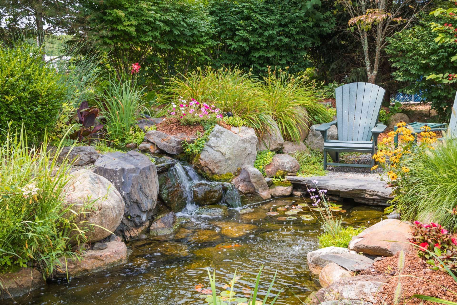 A small waterfall flows into a pond surrounded by lush plants, rocks, and a wooden Adirondack chair in a garden setting.