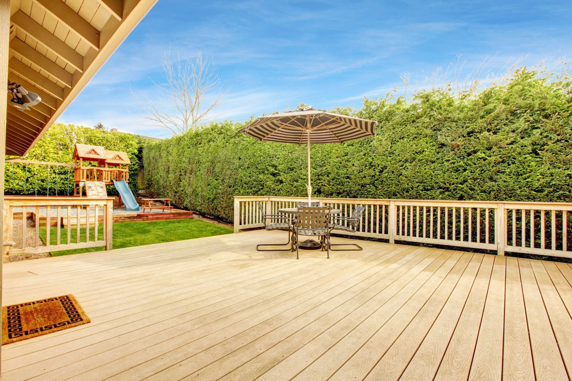 A wooden deck with a table, striped umbrella, and a railing, overlooking a green yard with a play set and tall hedge.