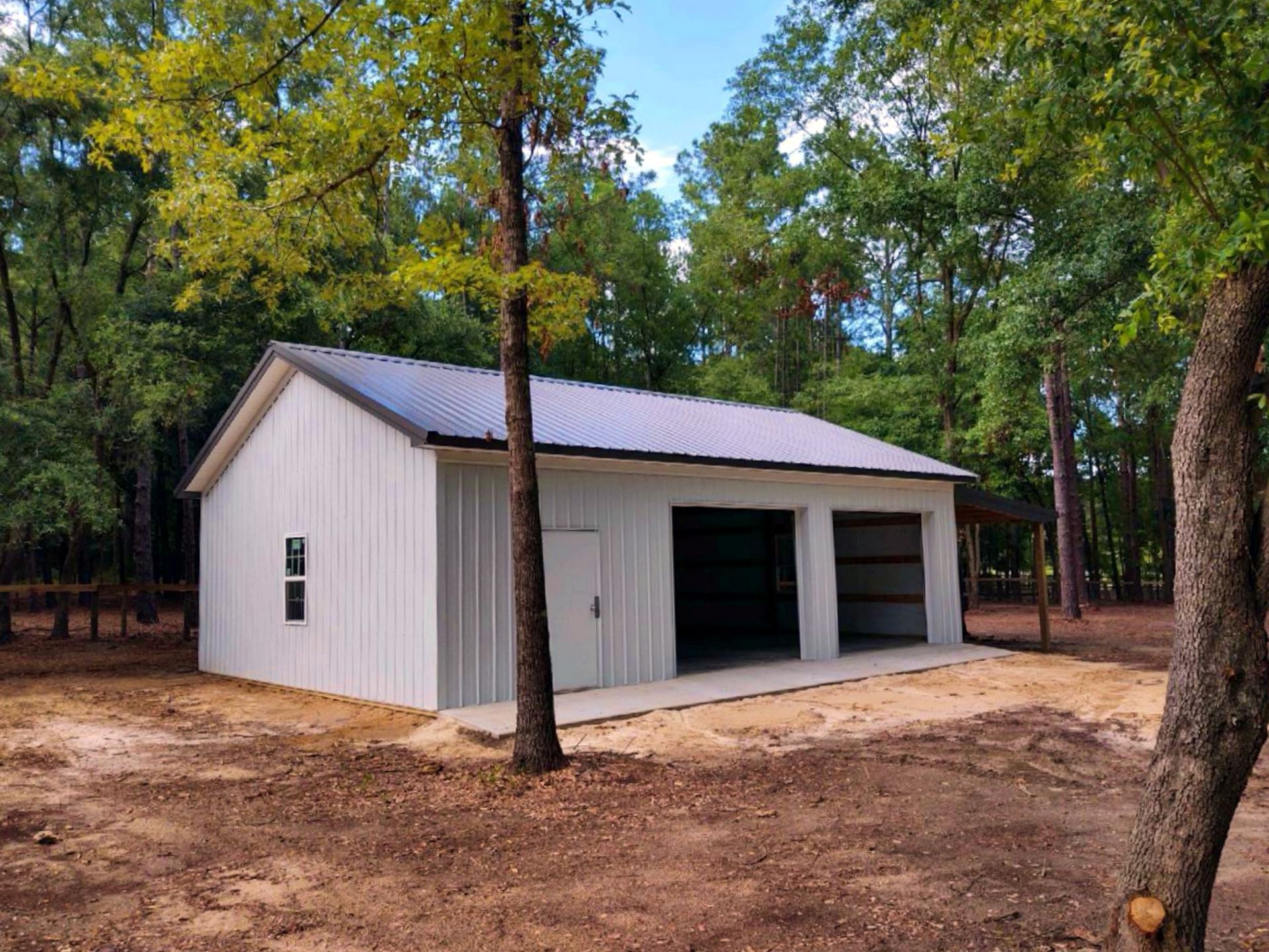 A white metal building with a grey roof and two open garage bays, situated in a wooded area on a dirt lot.