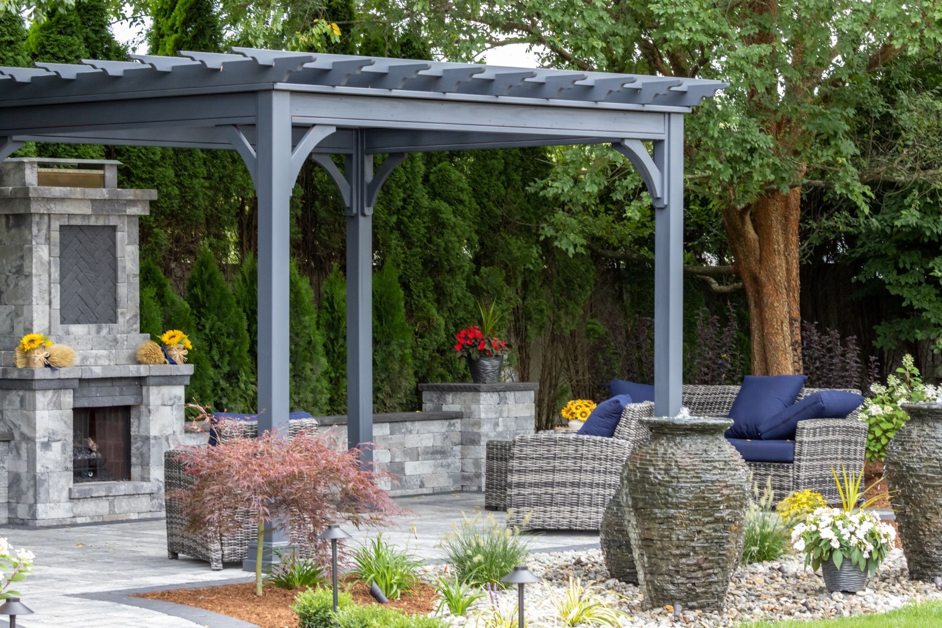 A gray wooden pergola over a stone fireplace and patio furniture with blue cushions in a lush backyard garden setting.