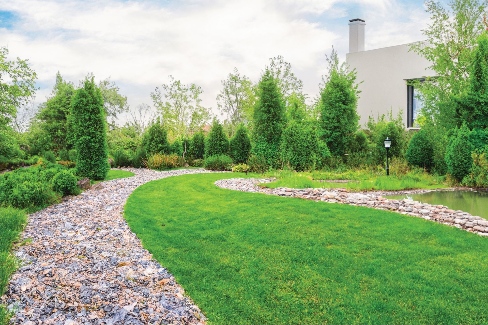 A stone pathway curves along a vibrant green lawn beside a calm pond, framed by evergreen trees and a modern home.