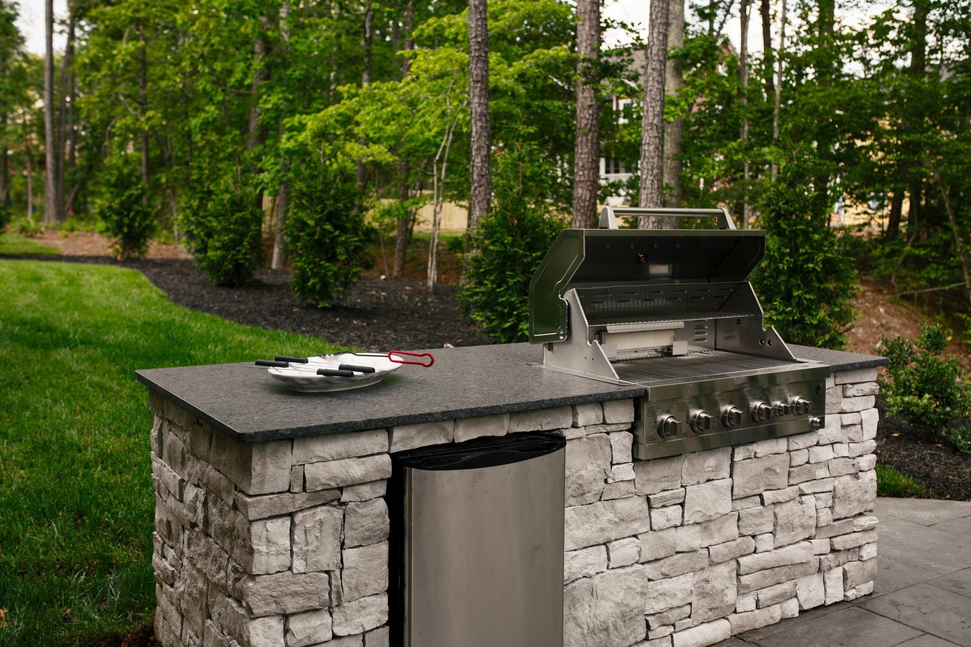 An outdoor stone kitchen island with a stainless steel grill and mini-fridge, set on a patio against a wooded backdrop.