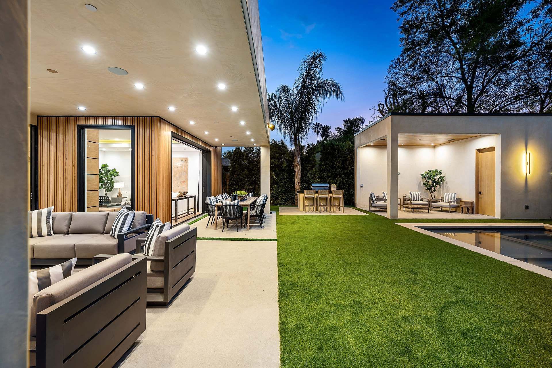 A modern patio with grey lounge furniture and a dining area overlooking a green lawn, swimming pool, and covered cabana.