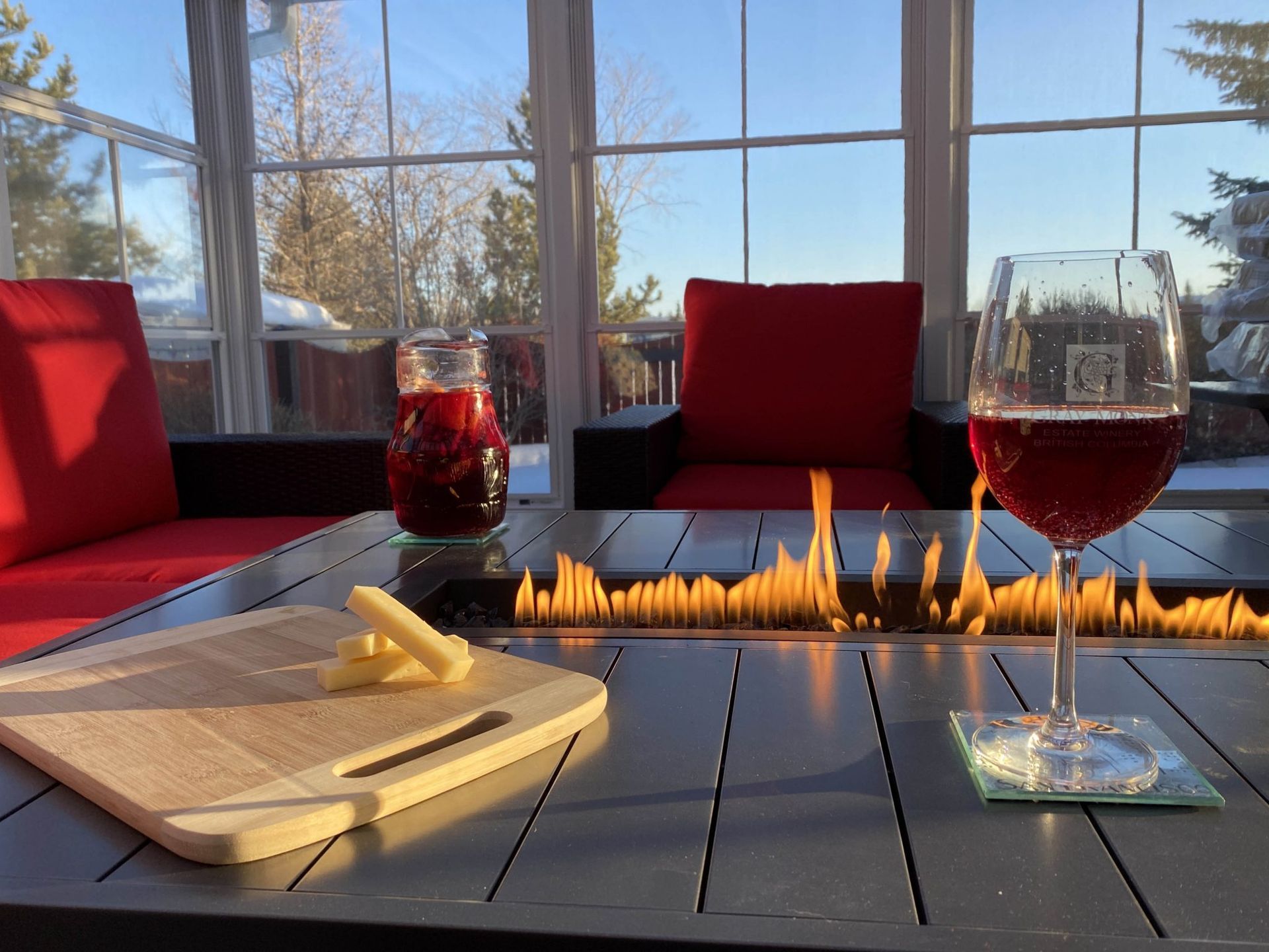Wine and candles on a sunlit patio table with red chairs and a window view