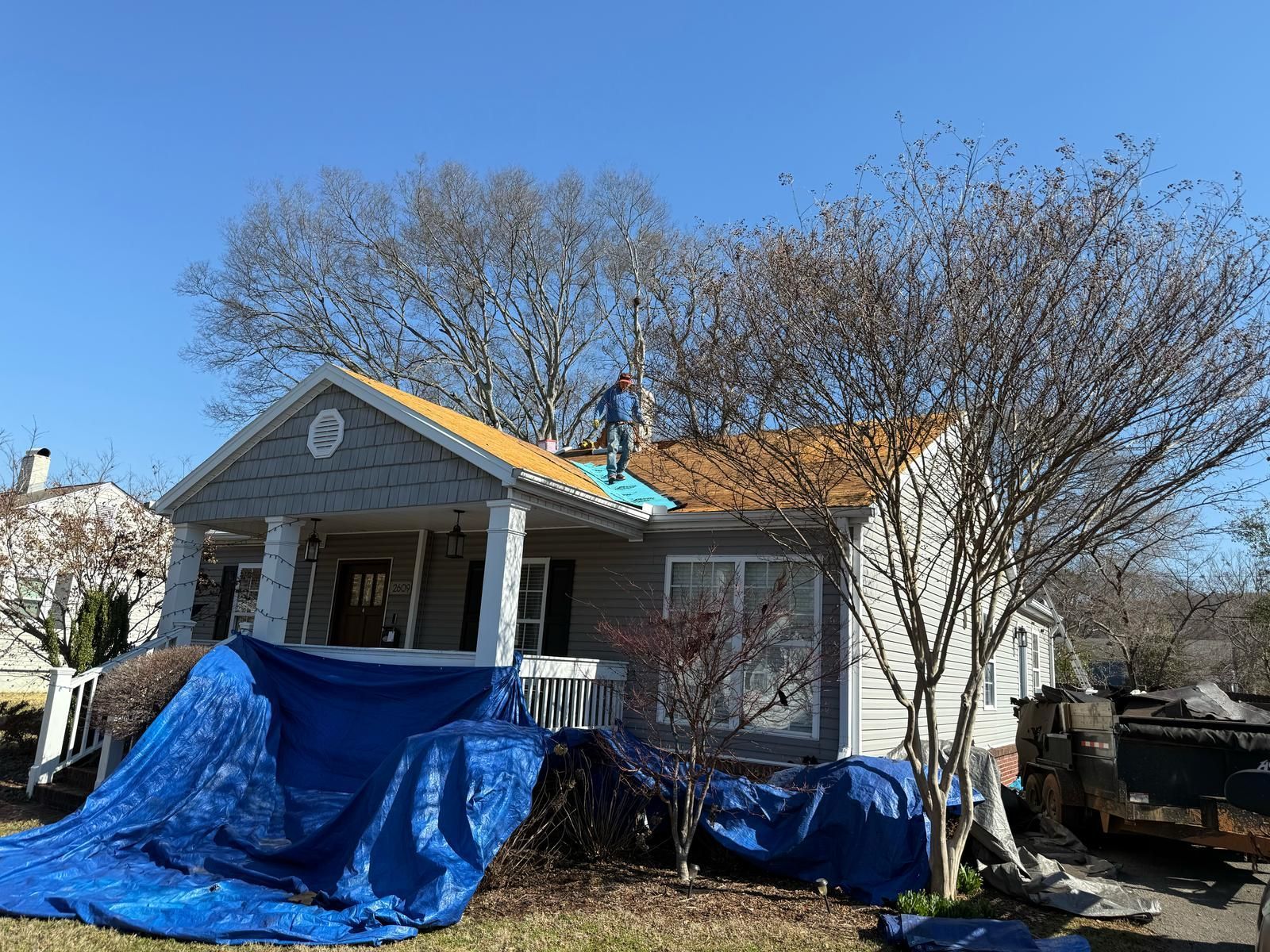 House with partially replaced roof; worker on roof, blue tarp covers porch.