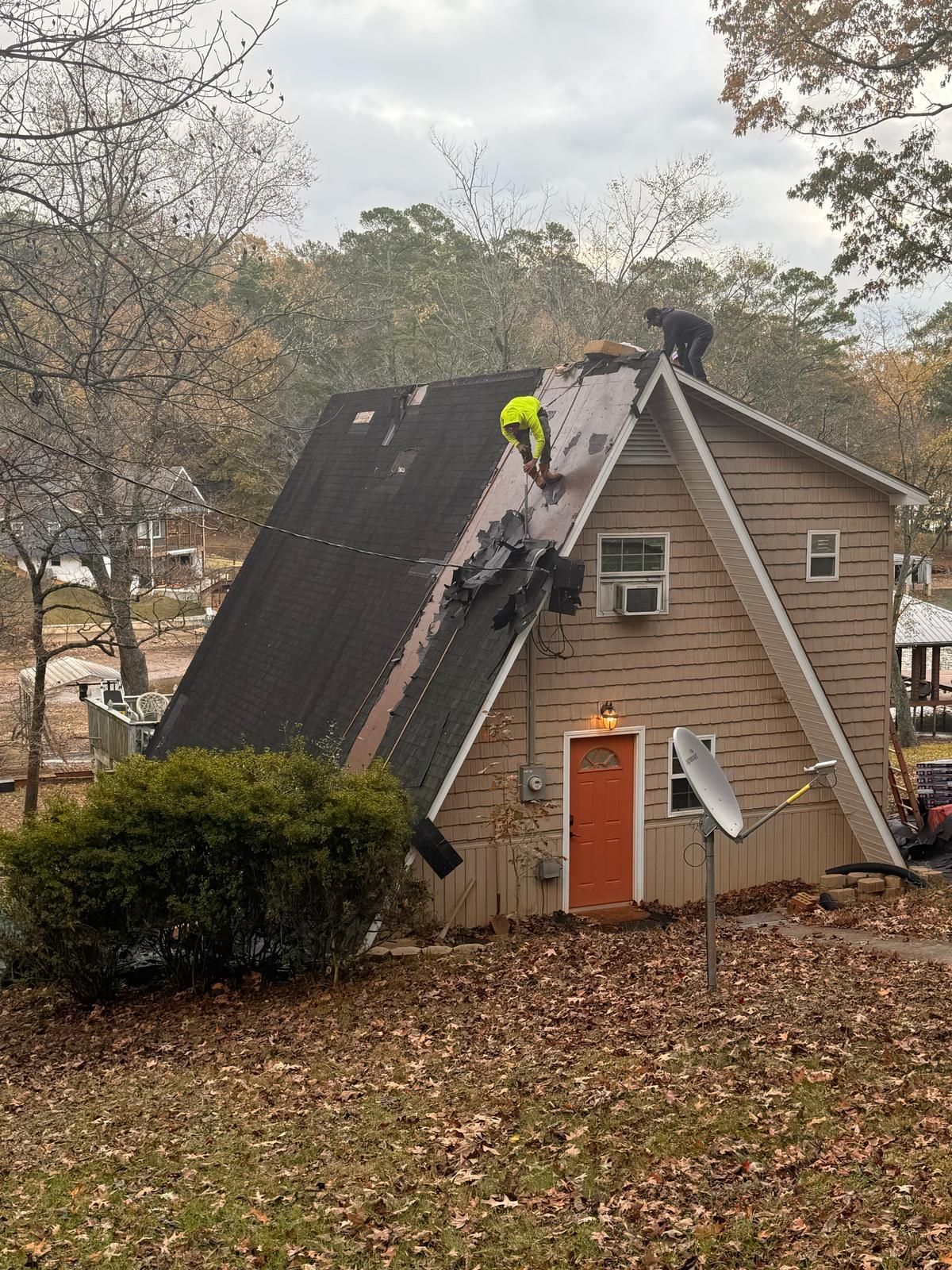 Two roofers replacing shingles on a brown A-frame house; overcast day, foliage in the background.