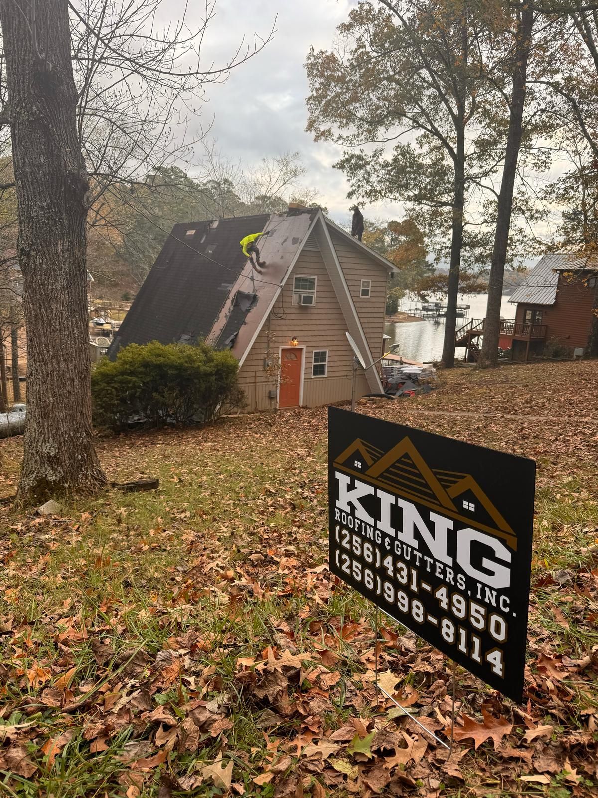 A-frame house with roofers, a lake in the background, and a King Roofing sign in the foreground.