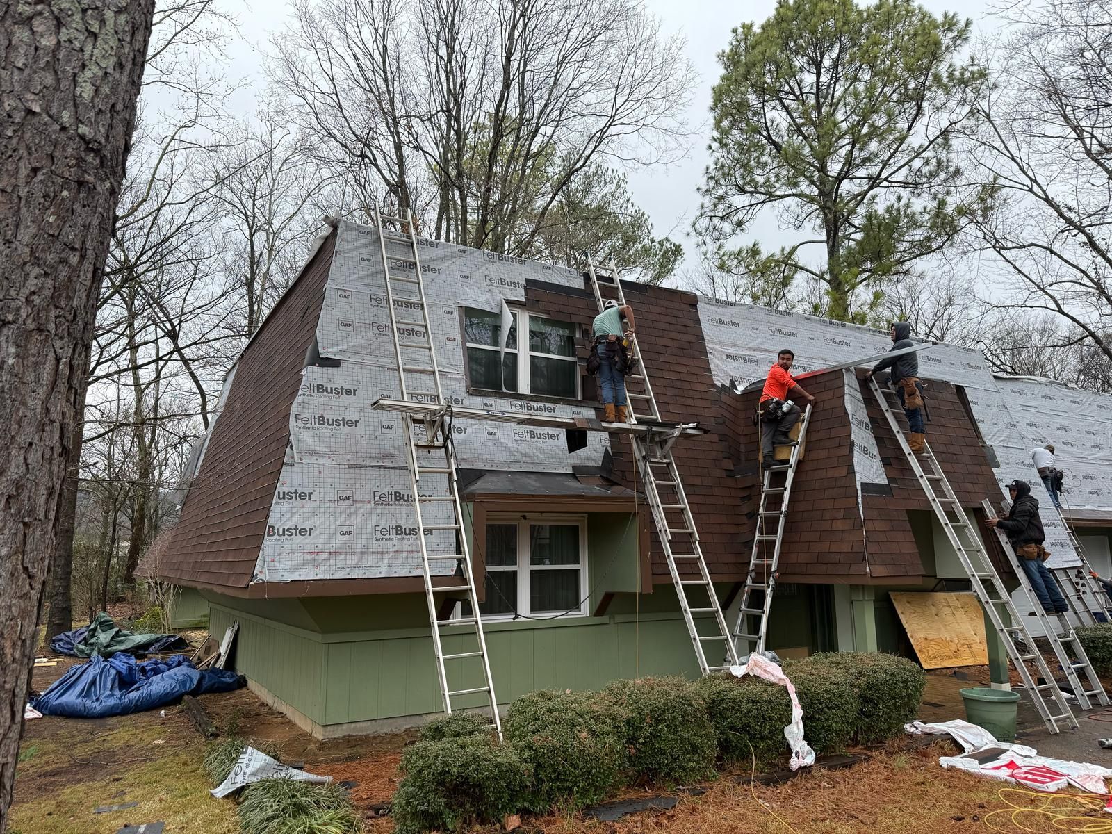 Roofers on ladders replacing shingles on a house with brown roofing. Overcast day.