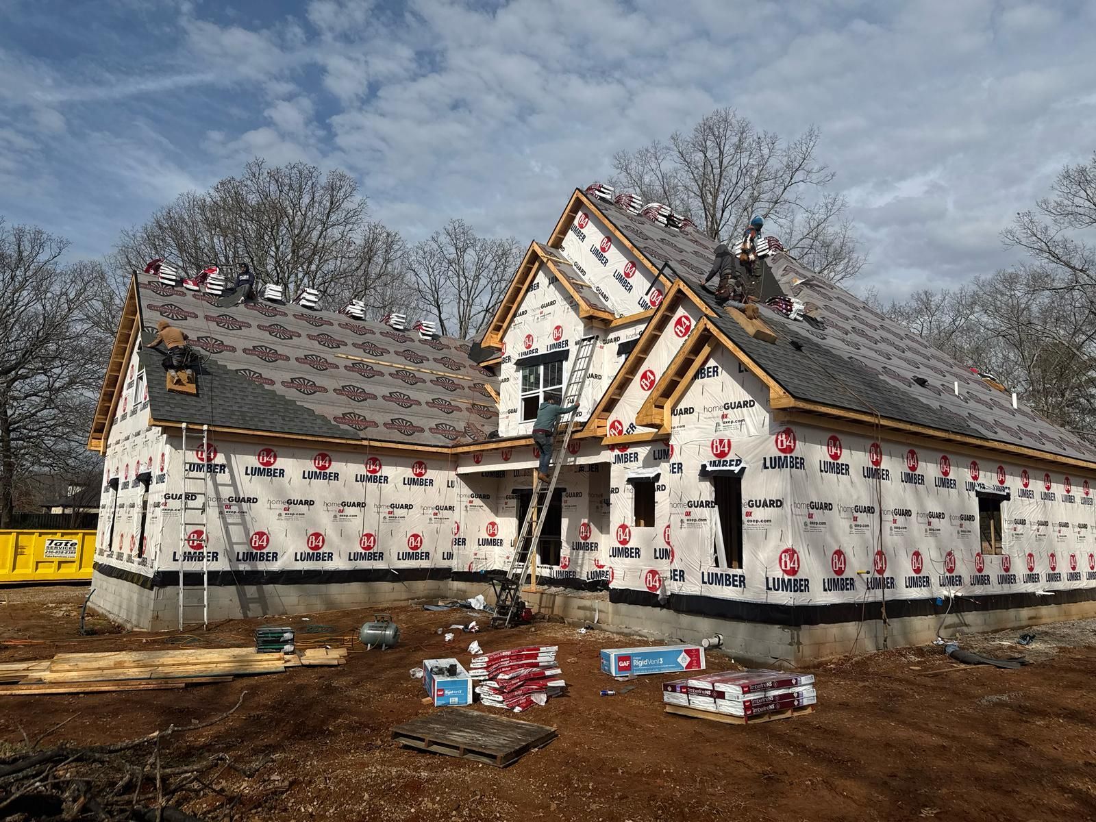 Construction site: House under construction, roofers working, building wrap visible, cloudy sky.