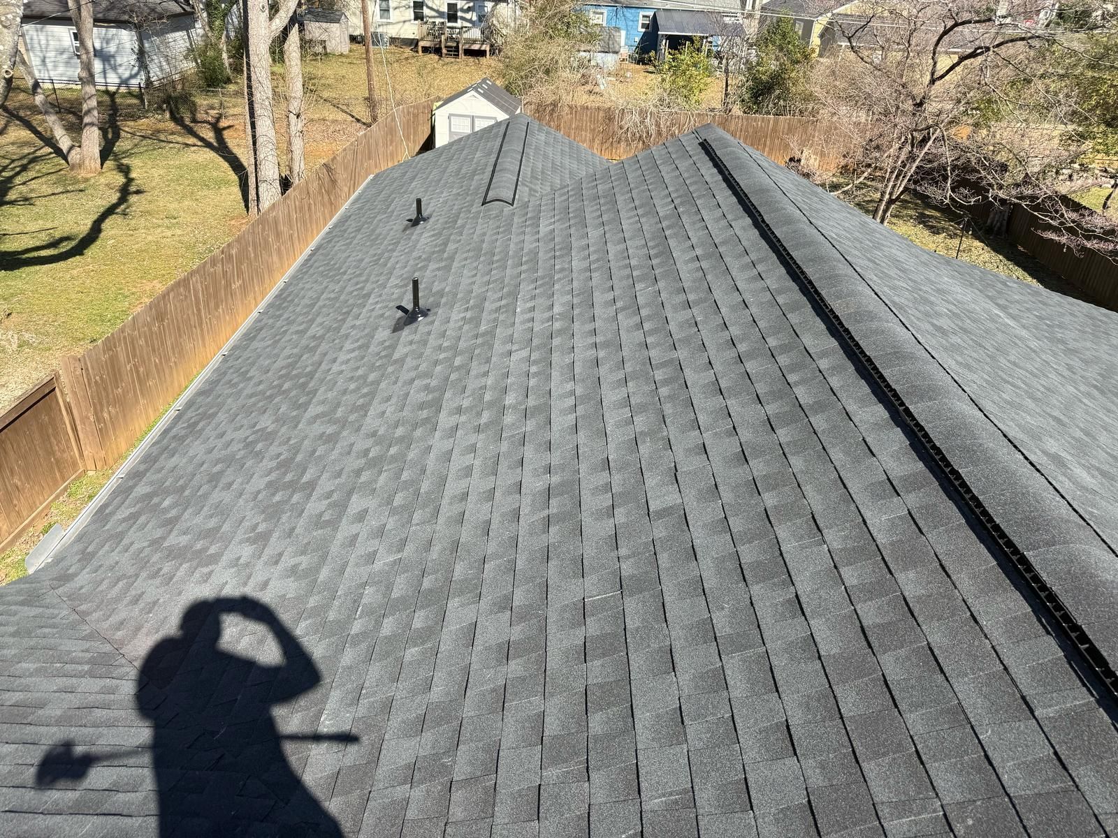 View of a dark gray asphalt shingle roof. A shadow of a person taking the photo is visible.