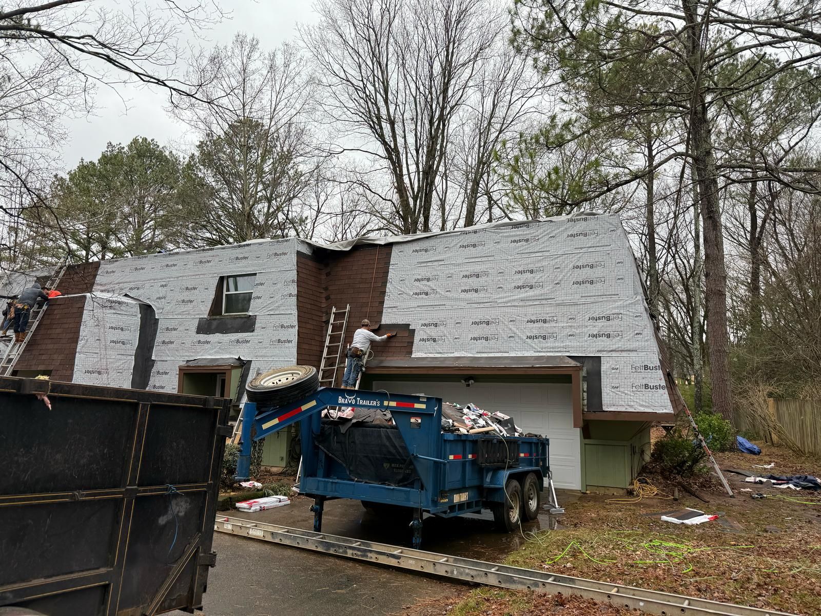 Workers replacing a roof on a house with a dumpster trailer parked in the driveway.