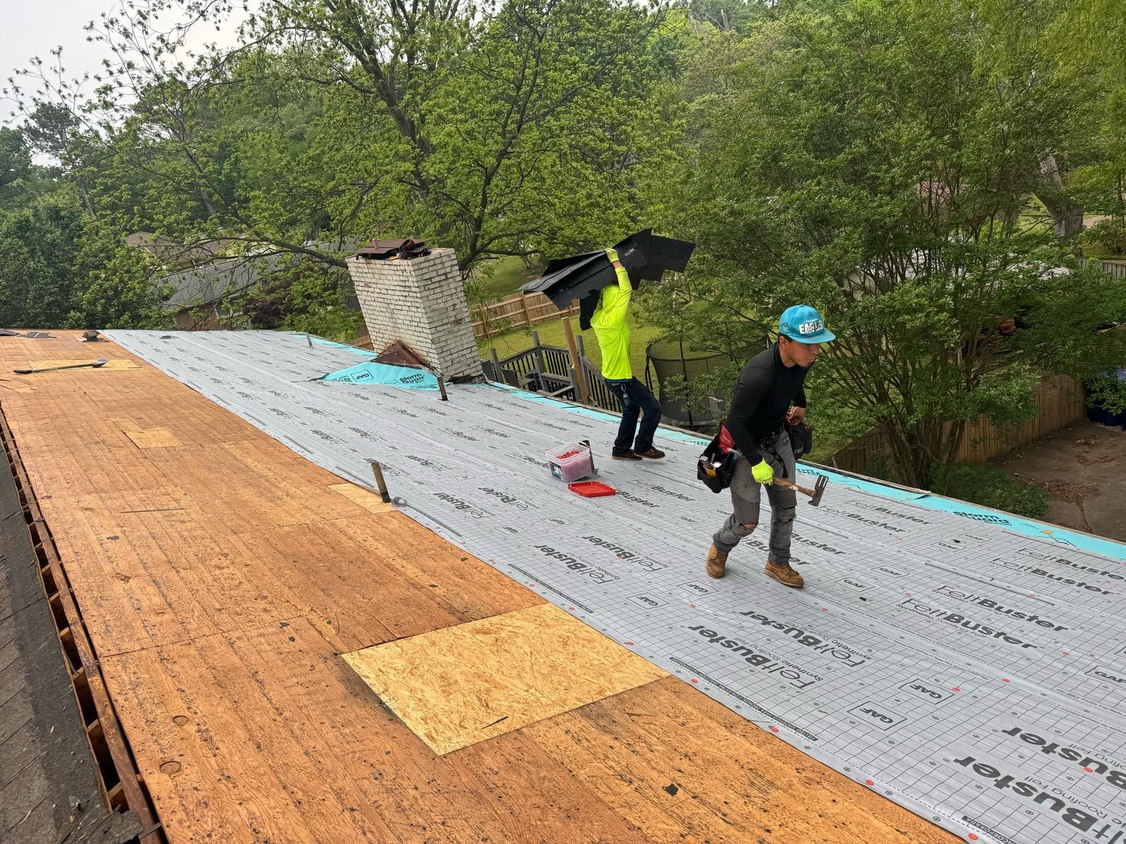 Two roofers work on a roof, installing materials. One hammers, the other carries shingles. Overcast day.