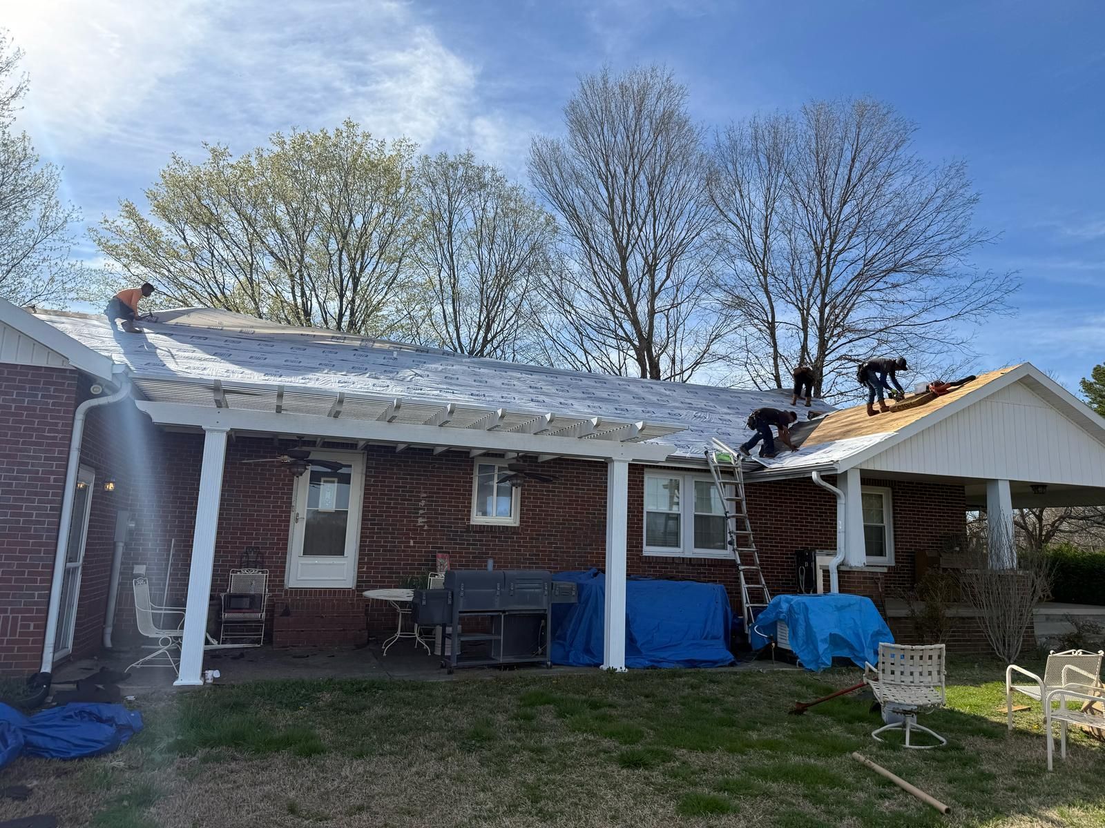 Workers on a roof, replacing shingles on a brick house. Ladders, tools, and tarps are visible.